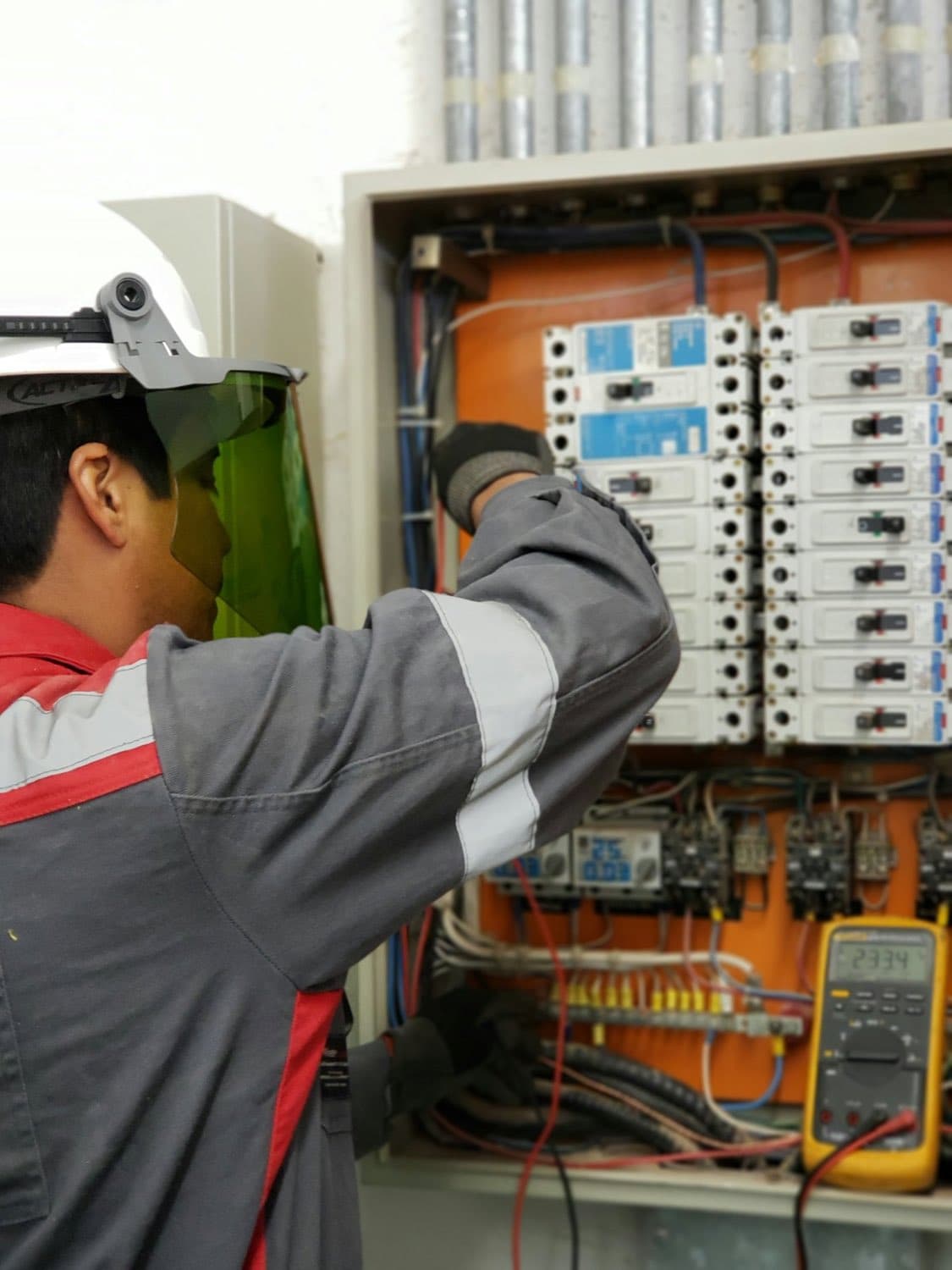 An electrician in a hard hat and face shield testing an electrical panel