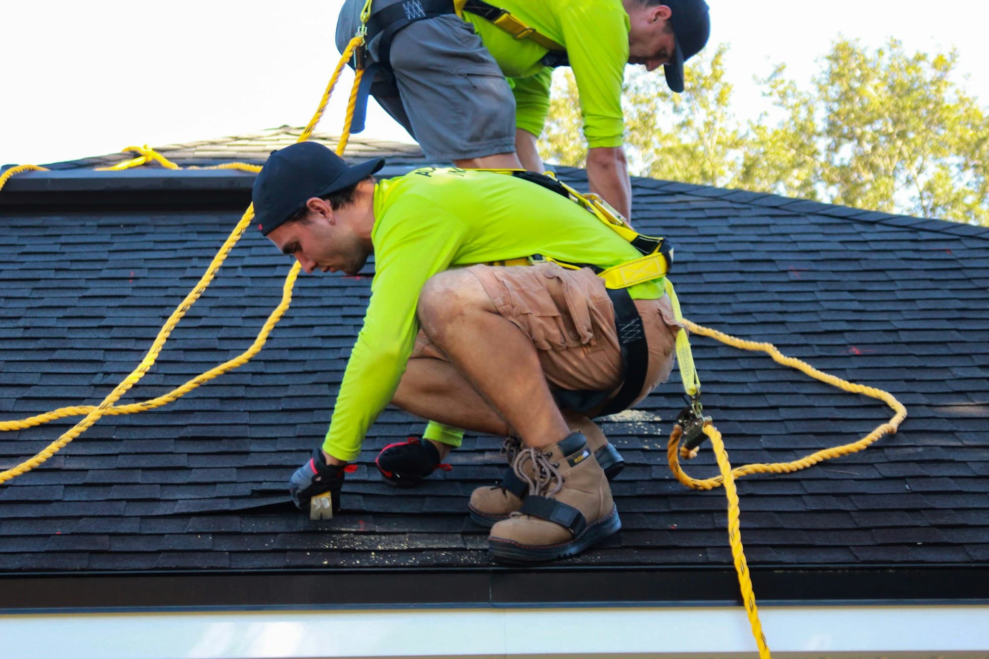 Two roofers in green shirts and safety harnesses working on dark shingles of a house roof