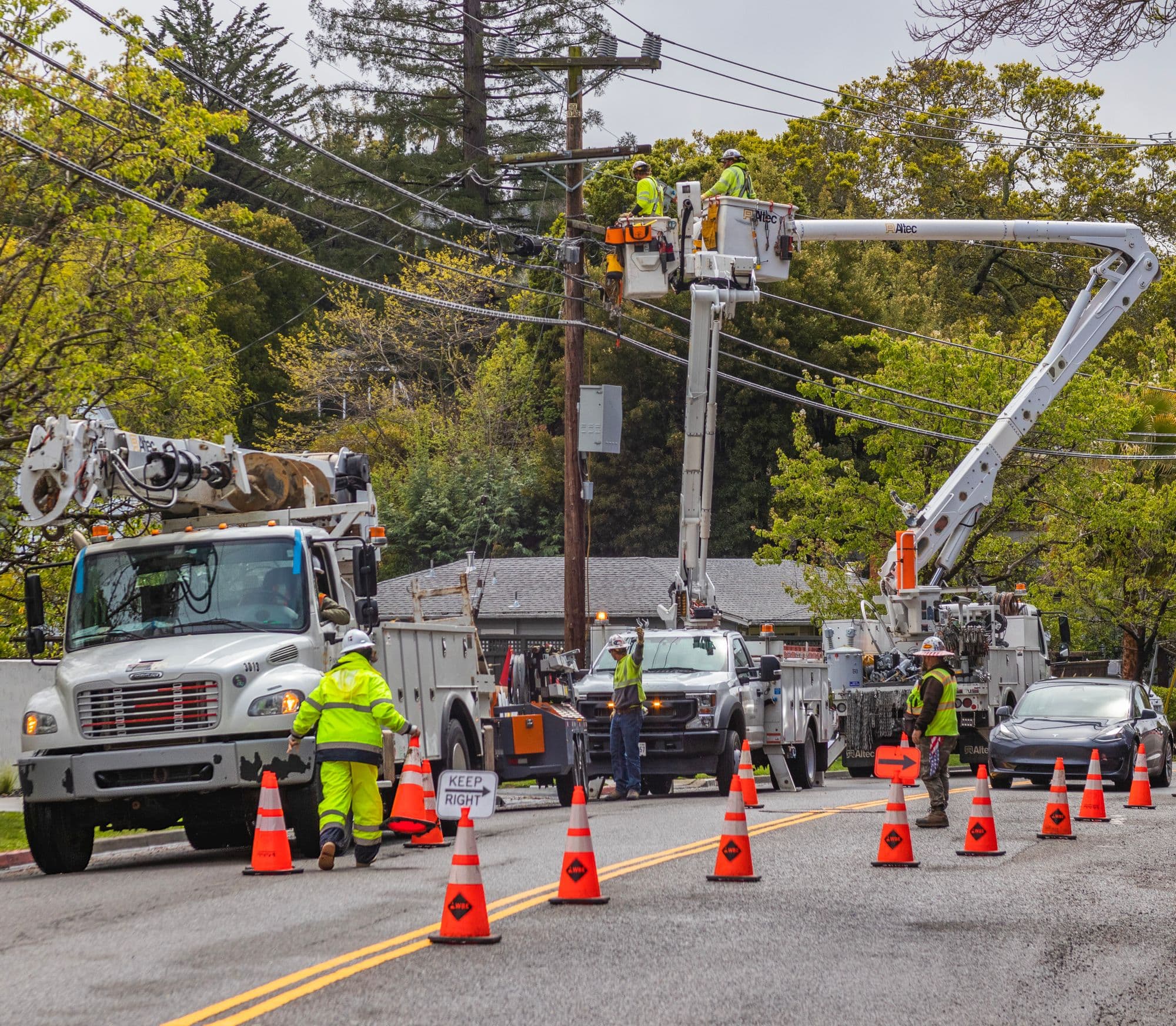 Utility crews in yellow safety vests work from two bucket trucks on a residential street, with traffic cones marking the lane