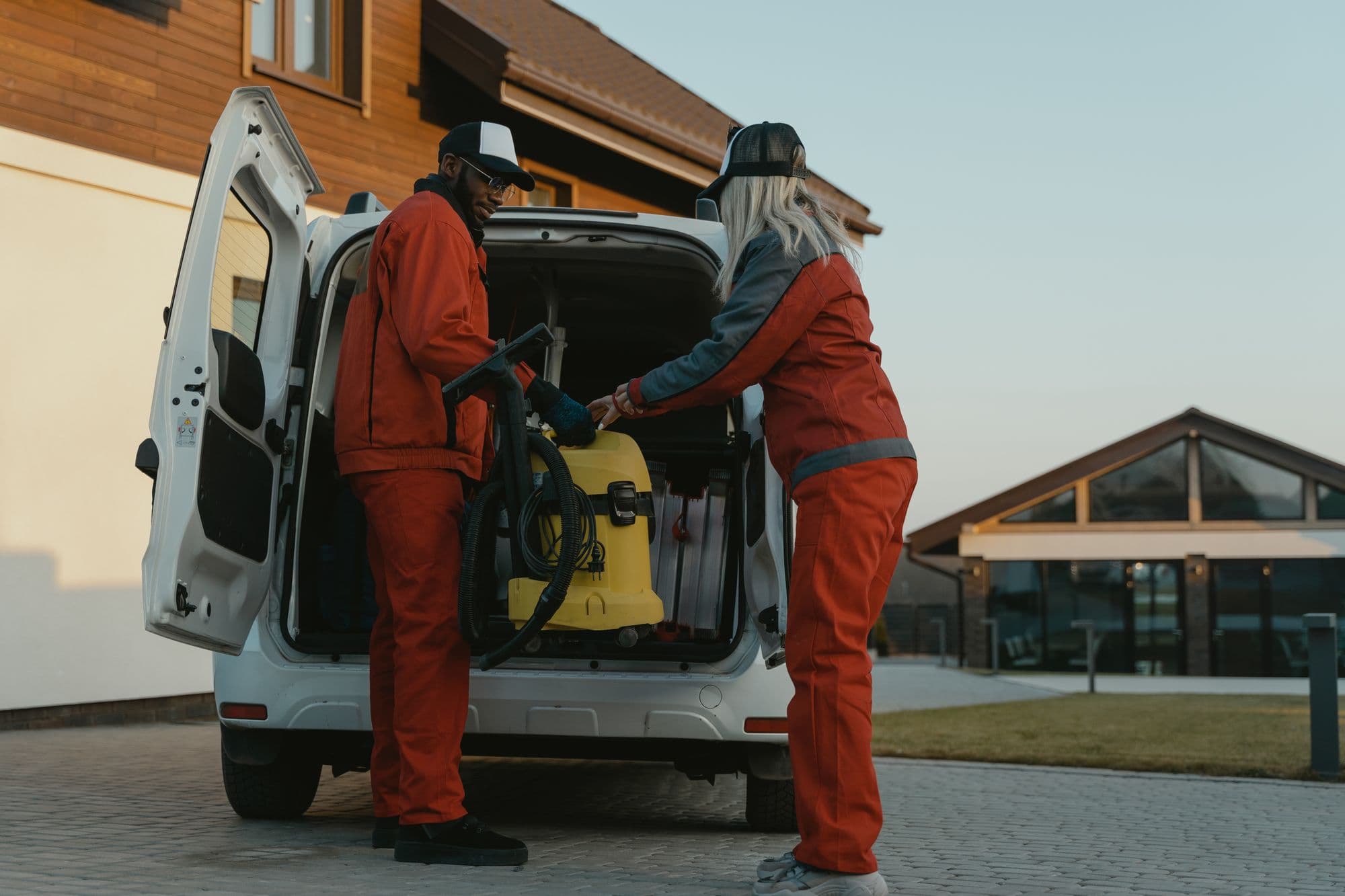 Two workers in orange uniforms and caps lifting a yellow shop vacuum out of the back of a white van in a driveway
