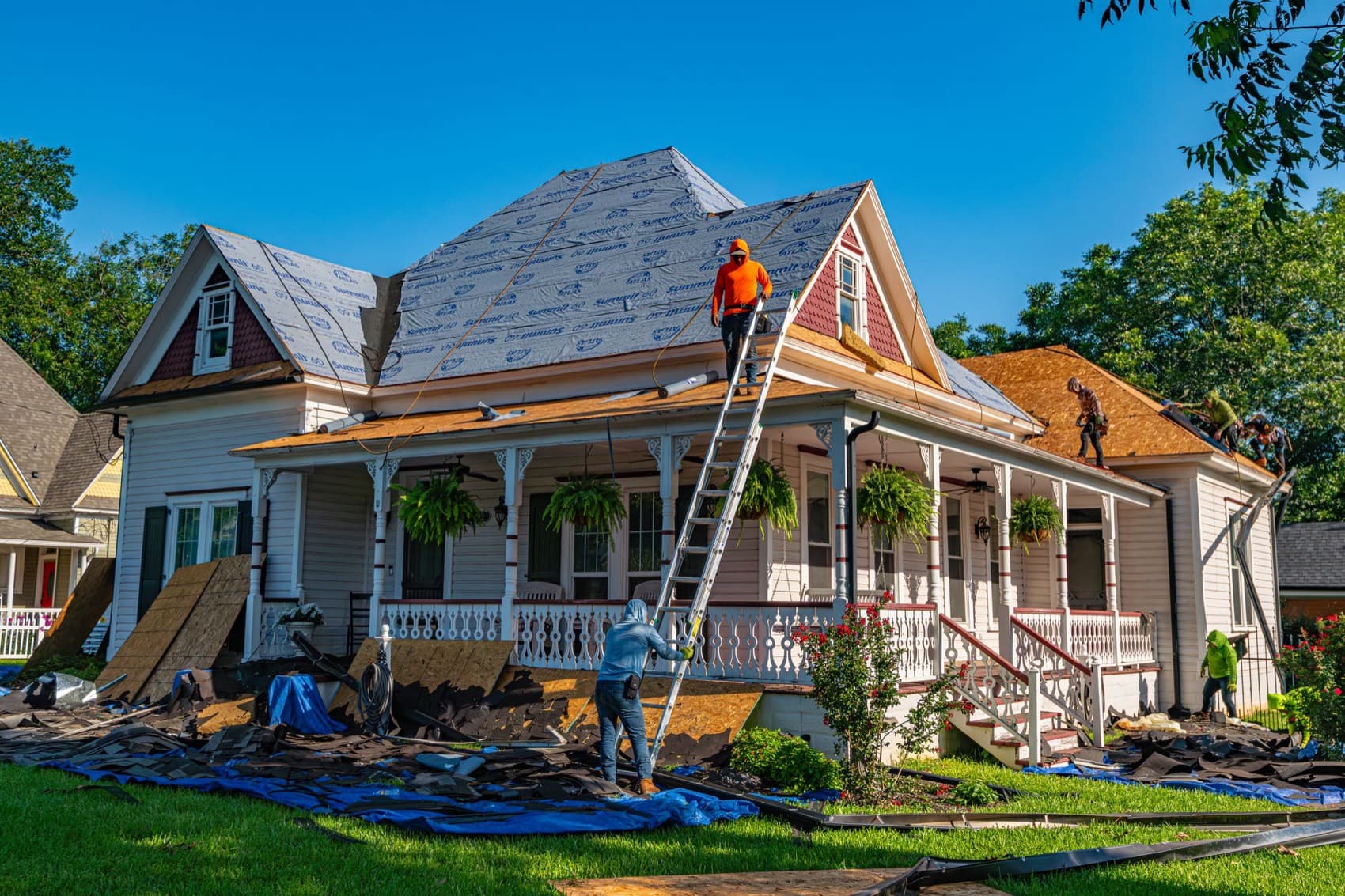 A roofing crew replacing the roof of a two-story house, ladders against the porch