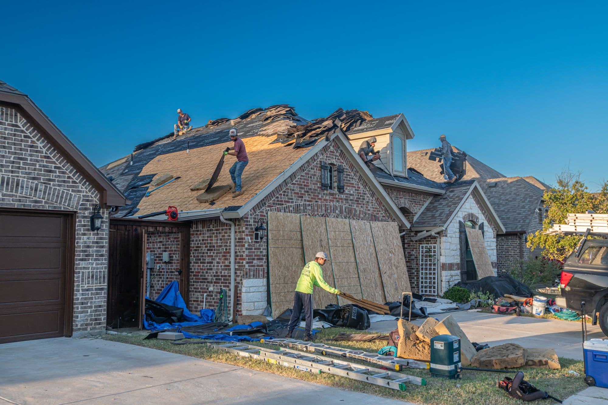 Roofers tearing off shingles on a brick house roof while a worker in a yellow shirt moves plywood panels below