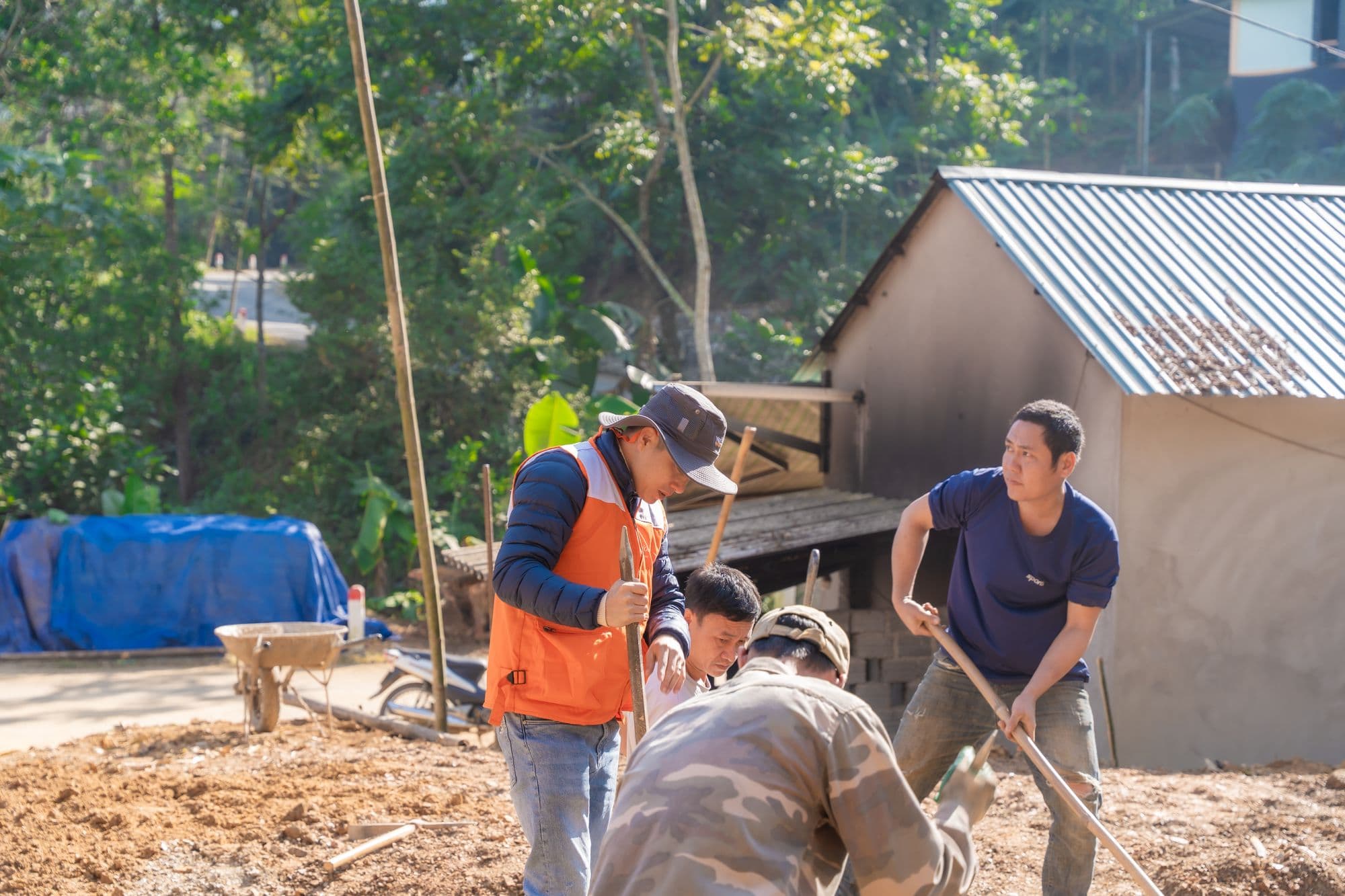 Four workers digging and raking gravel on a sunny job site beside a small house with a metal roof